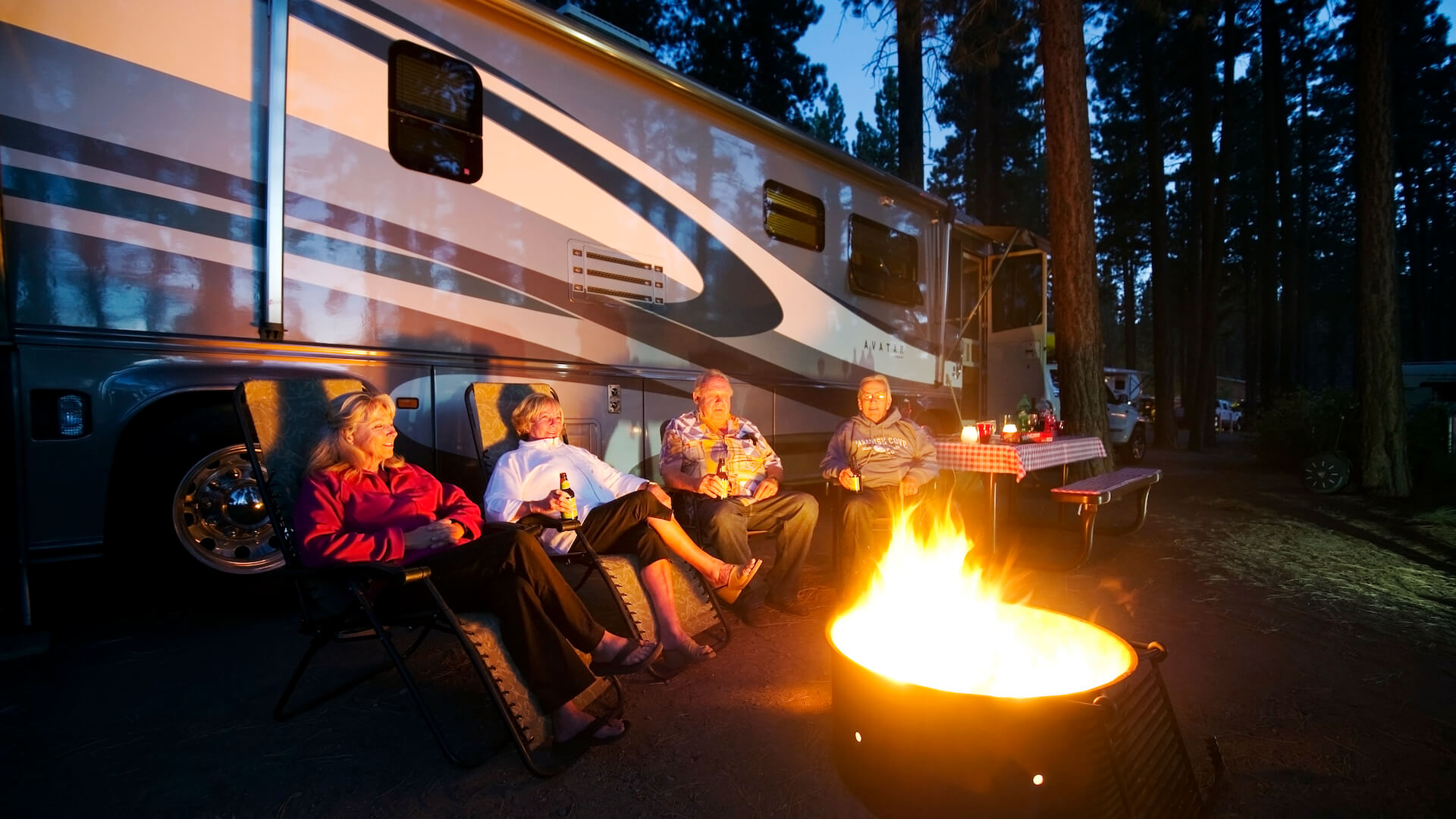 Four adults sitting between an RV and a camp fire